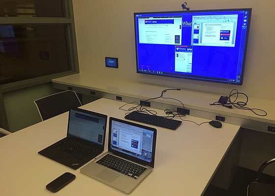 A conference room setup with two laptops, a smartphone, and a large wall-mounted screen displaying a presentation.